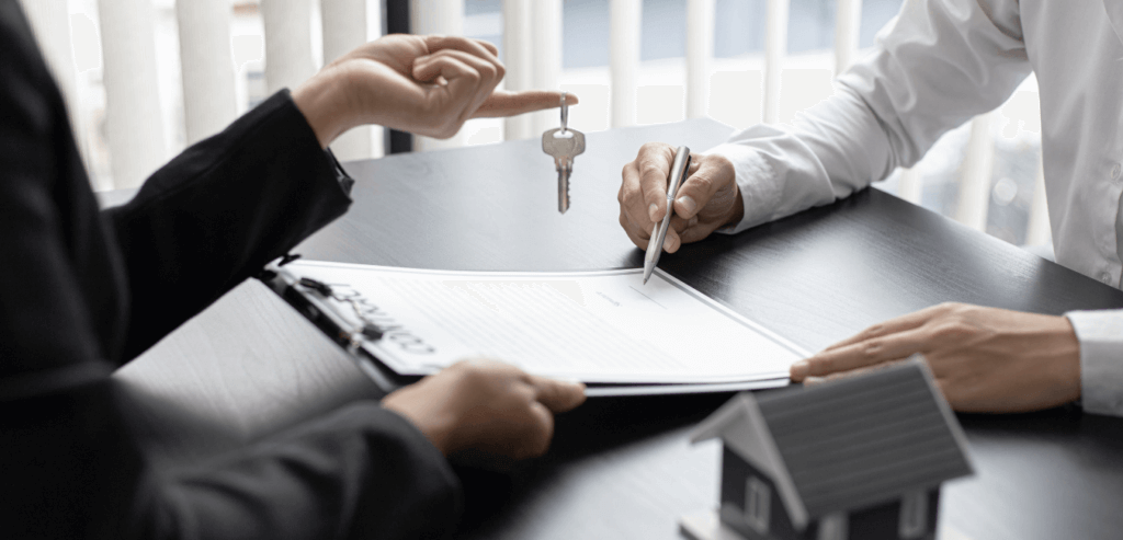 A picture of someone signing a document while another person holds a key over the paper.